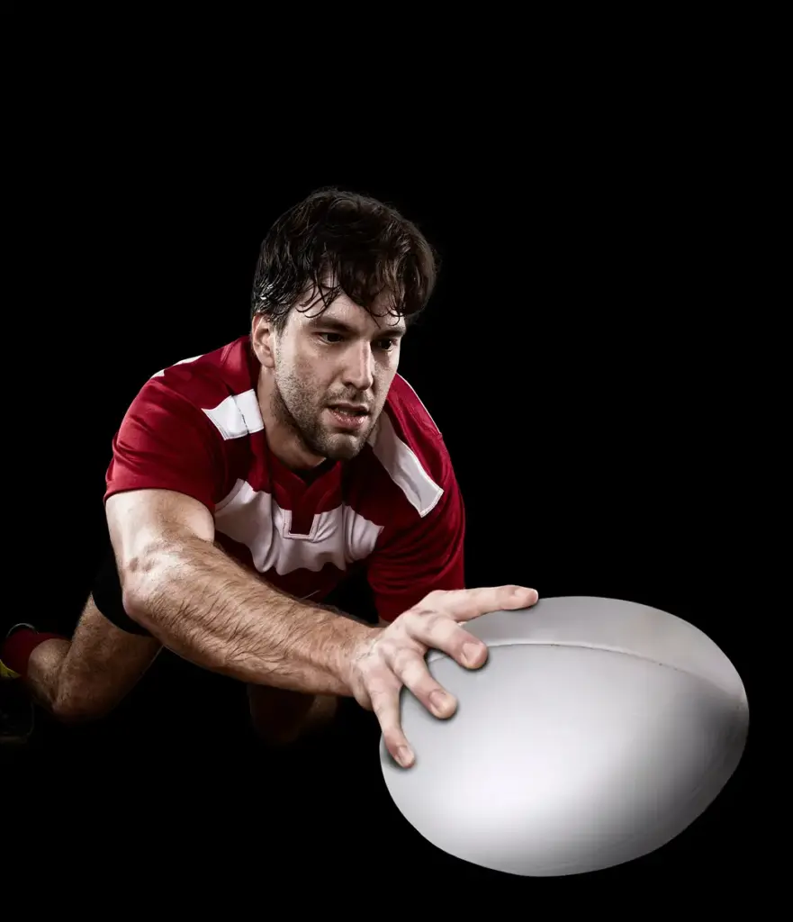 Um jogador profissional de râguebi, com um equipamento vermelho, lança-se para a frente enquanto estende o braço para colocar a bola no chão, tendo como pano de fundo um cenário escuro, captando um momento de ação dinâmico durante o jogo