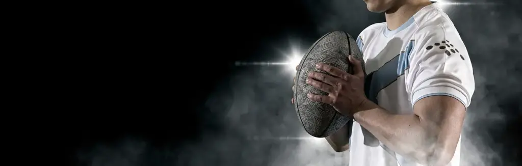 Um atleta de râguebi concentrado encontra-se sob a iluminação dramática do estádio, segurando uma bola de râguebi junto ao peito, criando uma atmosfera desportiva intensa e competitiva