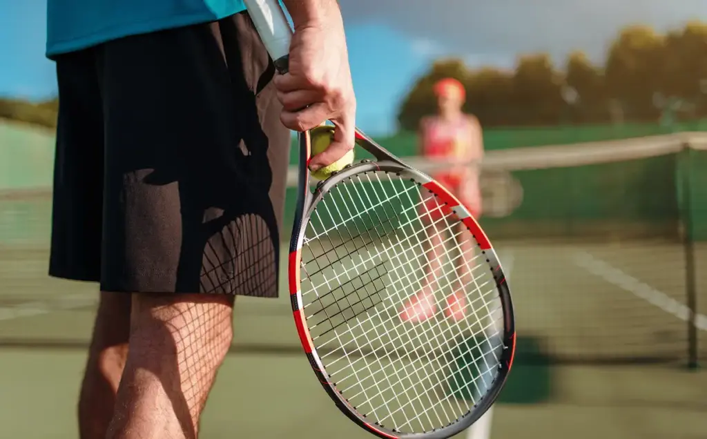 Um grande plano de um jogador de ténis segurando uma raquete e uma bola, em pé num campo de ténis ao ar livre, a preparar-se para o próximo serviço, com outro jogador desfocado ao fundo